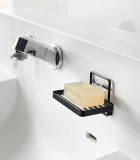 Yamazaki Home's black Traceless Adhesive Soap Tray mounted on a bathroom wall, holding a bar of yellow soap next to a chrome faucet. view 9