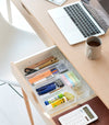 Three different sizes of stackable clear organizers—small, medium, and large—are neatly placed in a desk drawer. Made from transparent material, they accommodate stationery items like pens, paper clips, and sticky notes, showcasing their clarity and stackability.