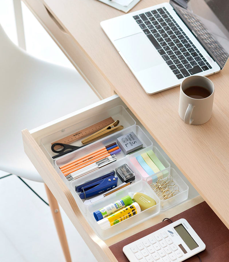 Three different sizes of stackable clear organizers—small, medium, and large—are neatly placed in a desk drawer. Made from transparent material, they accommodate stationery items like pens, paper clips, and sticky notes, showcasing their clarity and stackability.