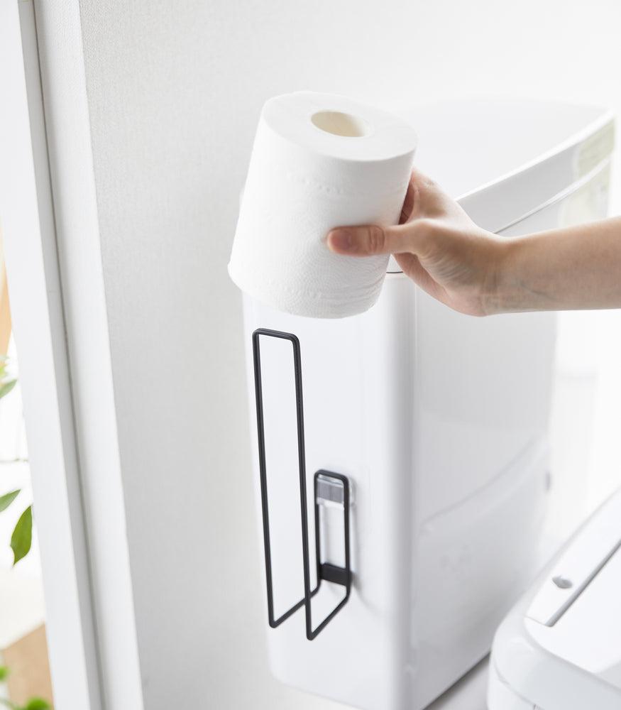 View 13 - A toilet paper roll being placed on a black Yamazaki Home Traceless Adhesive Toilet Paper Holder