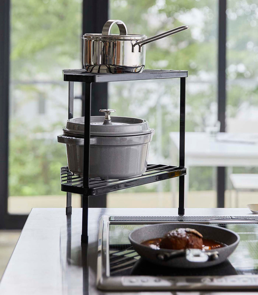 View 8 - Black Two-Tier Corner Riser holding pot and saucepan next to stovetop by Yamazaki Home.
