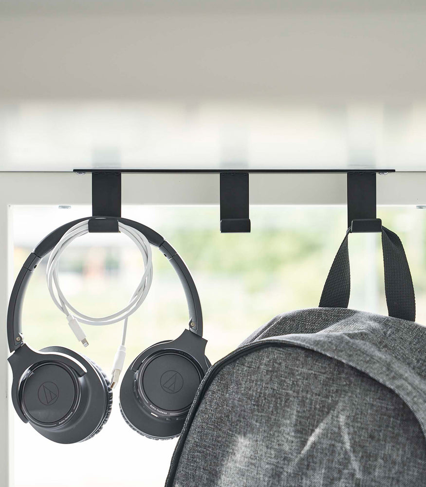 View 10 - Three matte black metal hooks are secured to the underside of white desk. One hook holds a pair of over-ear headphones and a wrapped charger cord, while another holds a backpack.