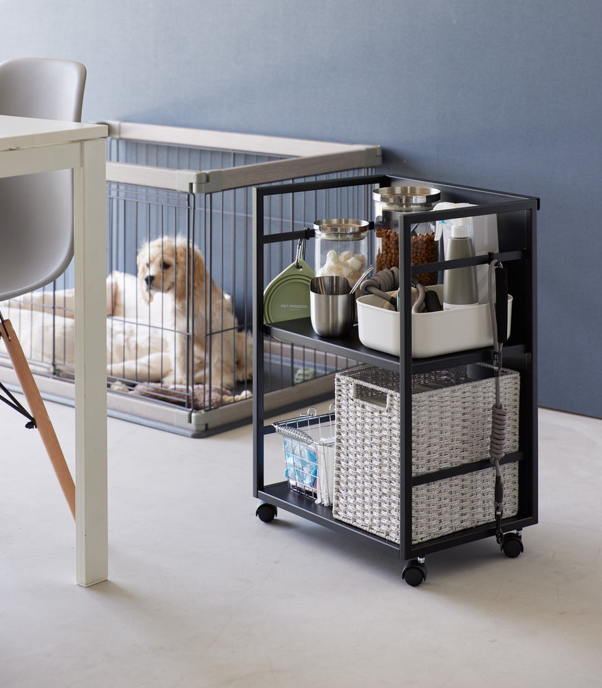 View 12 - Two-level black steel storage cart with casters is shown next to a dog crate. Cart houses various dog supplies and treat storage on the upper shelf, bottom shelf holds two storage bins. Hooks are installed on both sides, one holds a collapsible water bowl, the other a leash. Solid back wall of the cart demonstrates that the items can be hidden when flipped.