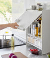 Image showing a hand lifting the front panel of the Concealable Spice Rack by Yamazaki Home in white by the handle, revealing various condiments inside.