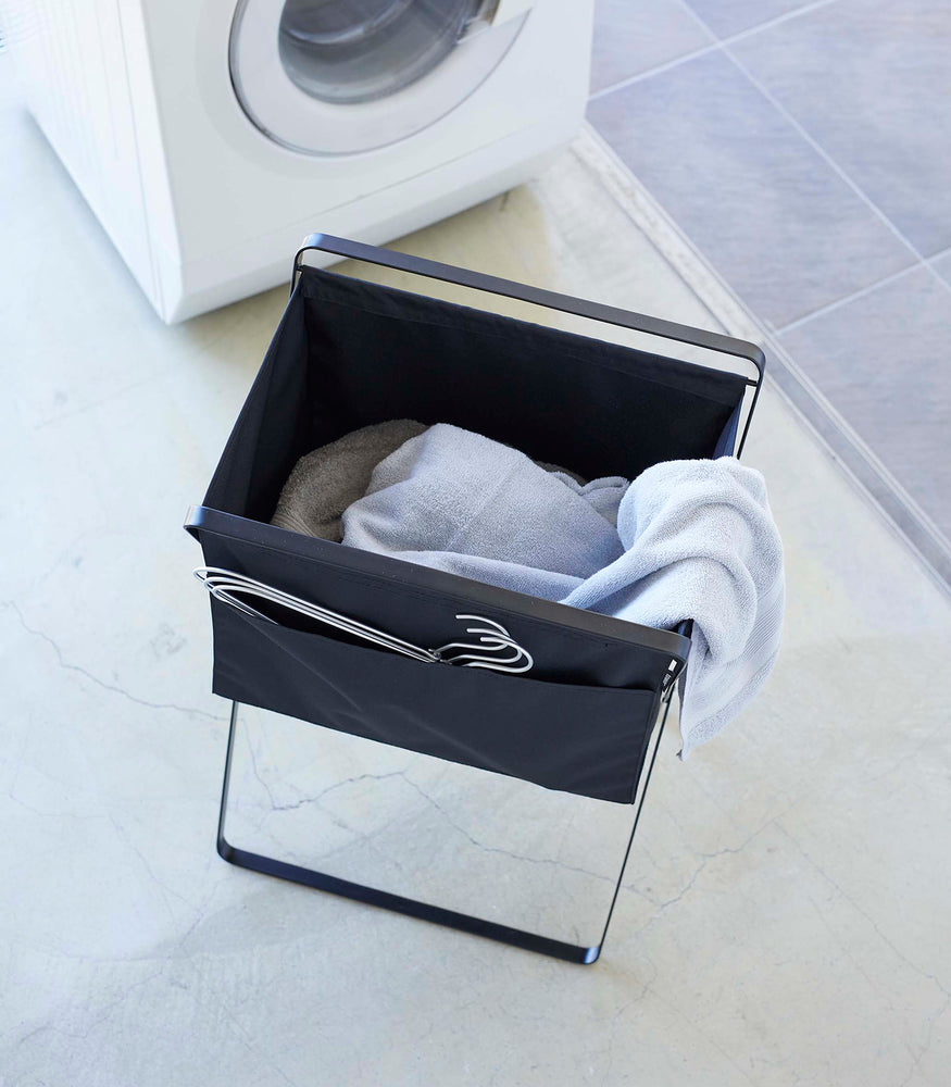 View 27 - Bird’s eye view of a black canvas hamper with multiple towels inside and wired hangers peeking out of a side-pocket. A washing machine is seen in the background.