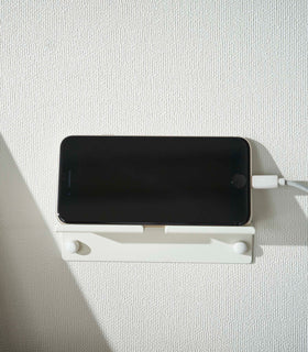 An iPhone with a charger cord plugged in is mounted on a wall above a desk setup. The screen is facing outward and the phone is held up on its side. view 5