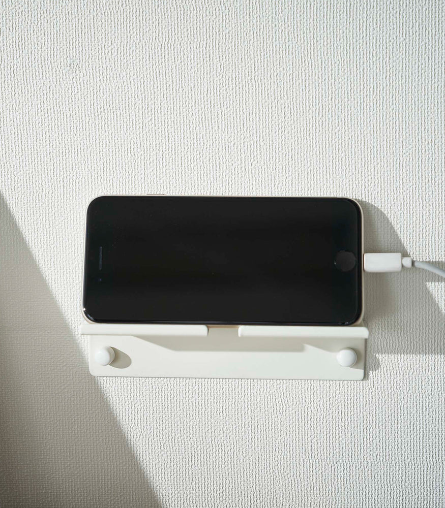 View 5 - An iPhone with a charger cord plugged in is mounted on a wall above a desk setup. The screen is facing outward and the phone is held up on its side.