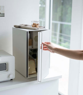 A vertical white metal breadbox is seen on a white kitchen counter next to a white microwave oven. The breadbox’s door is being swung opened by a person with only the arm in view. The door is opening from the right. A magnetic stop is seen opposite the open door. On top of the box is a folded towel and plastic container of cookies. view 16