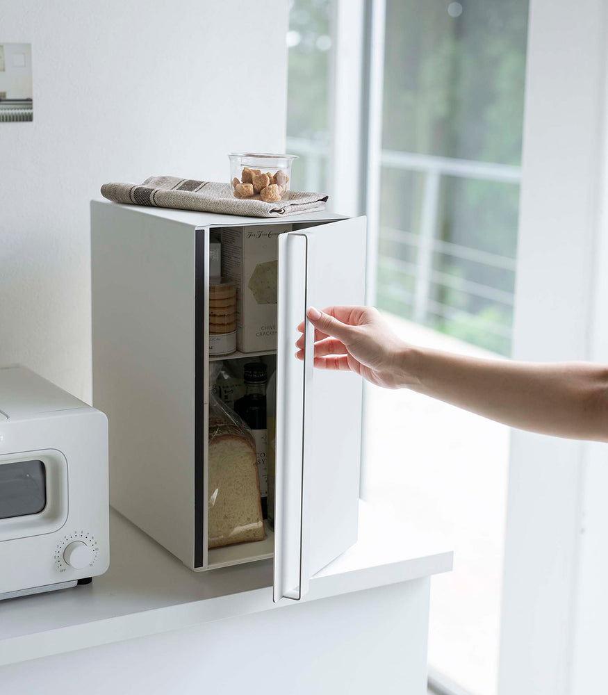 View 16 - A vertical white metal breadbox is seen on a white kitchen counter next to a white microwave oven. The breadbox’s door is being swung opened by a person with only the arm in view. The door is opening from the right. A magnetic stop is seen opposite the open door. On top of the box is a folded towel and plastic container of cookies.