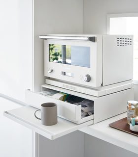 Diagonal view of the Countertop Drawer with Pull-Out Shelf by Yamazaki Home in white with the drawer and shelf pulled out. A mug sits on the shelf and a small microwave sits atop the unit. view 2