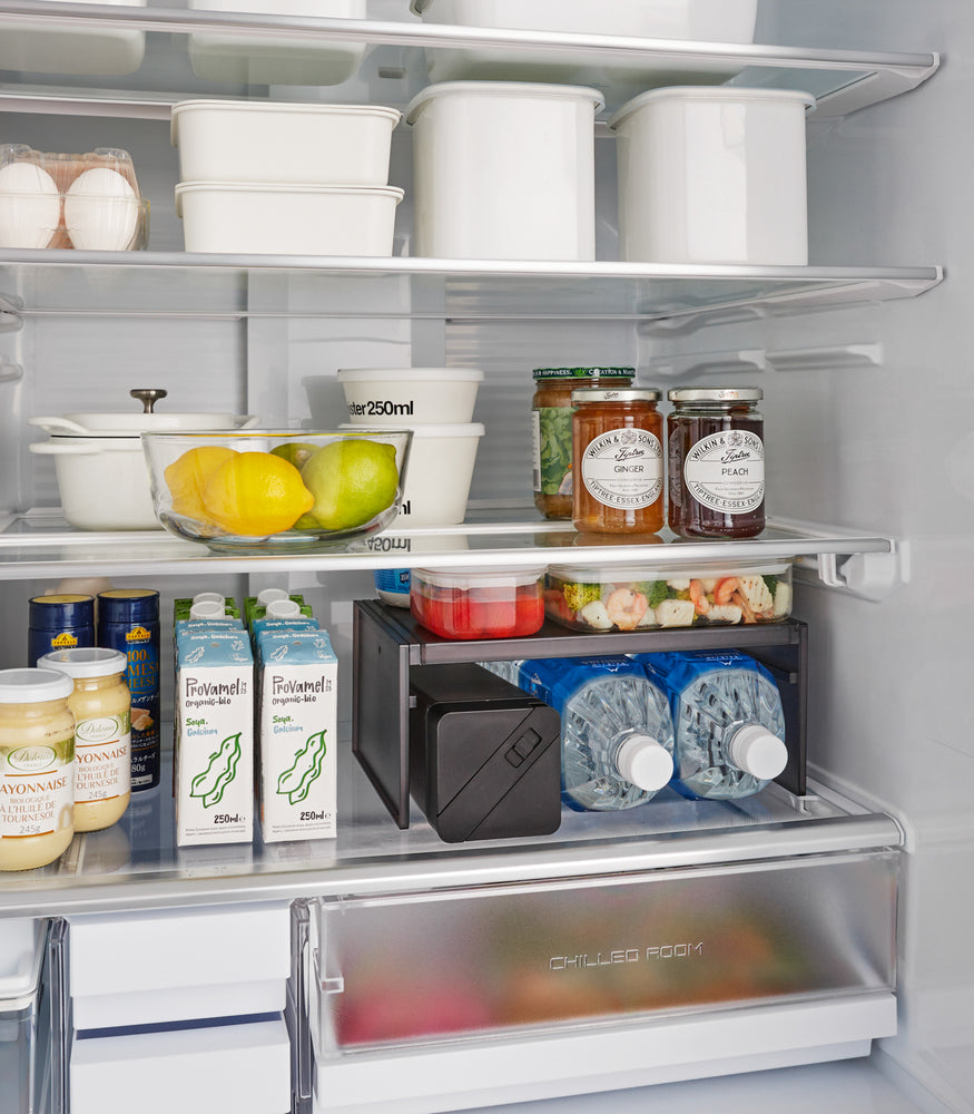 View 9 - A Black Expandable Refrigerator Riser Shelf by Yamazaki Home displayed in an open fridge, holding food items, demonstrating space utilization.