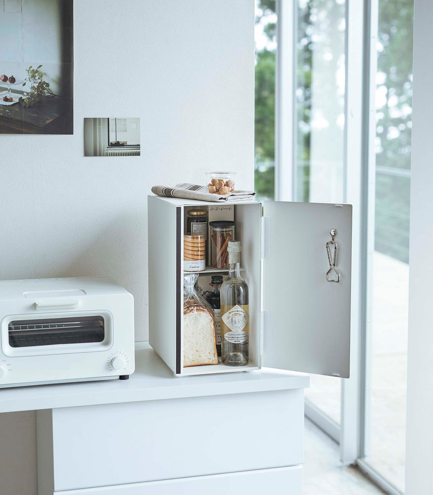 View 14 - A vertical white metal breadbox is seen on a white kitchen counter next to a white microwave oven and a glass drip coffee pot with a dark liquid inside.