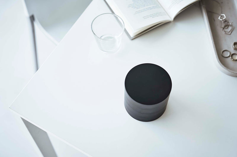 A bird’s-eye view of the corner of a white table. On the table is a cylinder-shaped black four-tier swivel accessory holder, a clear drinking glass, and an opened book. The accessory holder’s tiers and lid are swiveled closed so the inside contents cannot be seen, resembling a black cylinder object.