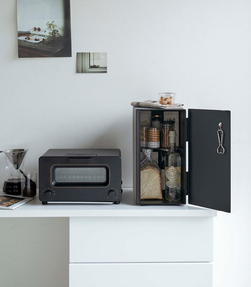 View 22 - A vertical black metal breadbox is seen on a white kitchen counter next to a black microwave oven and a glass drip coffee pot with a dark liquid inside.