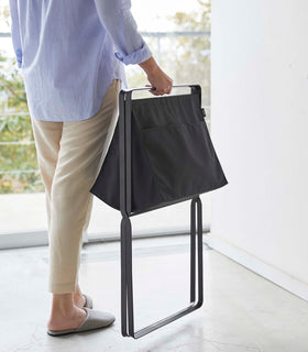 A person is walking toward a large window carrying a black canvas hamper by its folded black metal legs. view 14