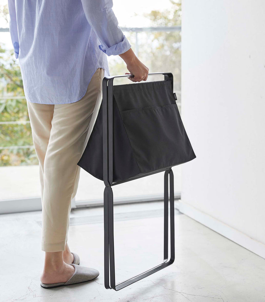 View 26 - A person is walking toward a large window carrying a black canvas hamper by its folded black metal legs.