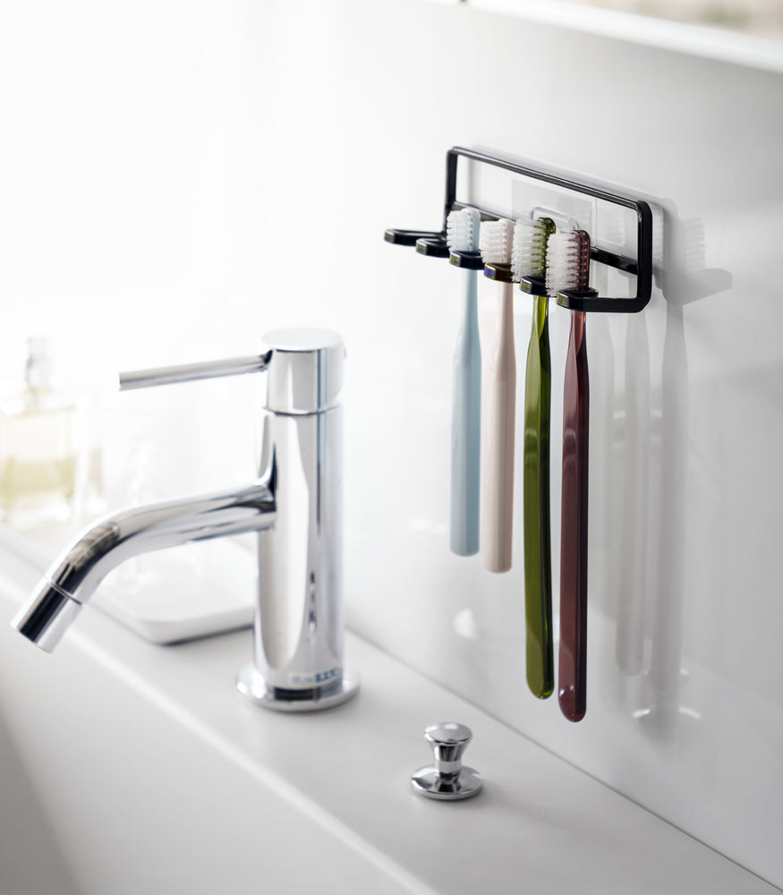 View 7 - Yamazaki Home's Traceless Adhesive Toothbrush Holder, black, mounted on a bathroom wall with four colorful toothbrushes and a chrome faucet.