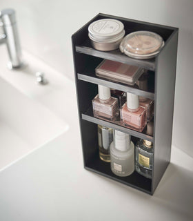 Angled view of a black rectangular cosmetics organizer sitting on a white bathroom counter. The organizer has three black transparent shelves with upward facing lips to prevent the products from falling-out. view 32