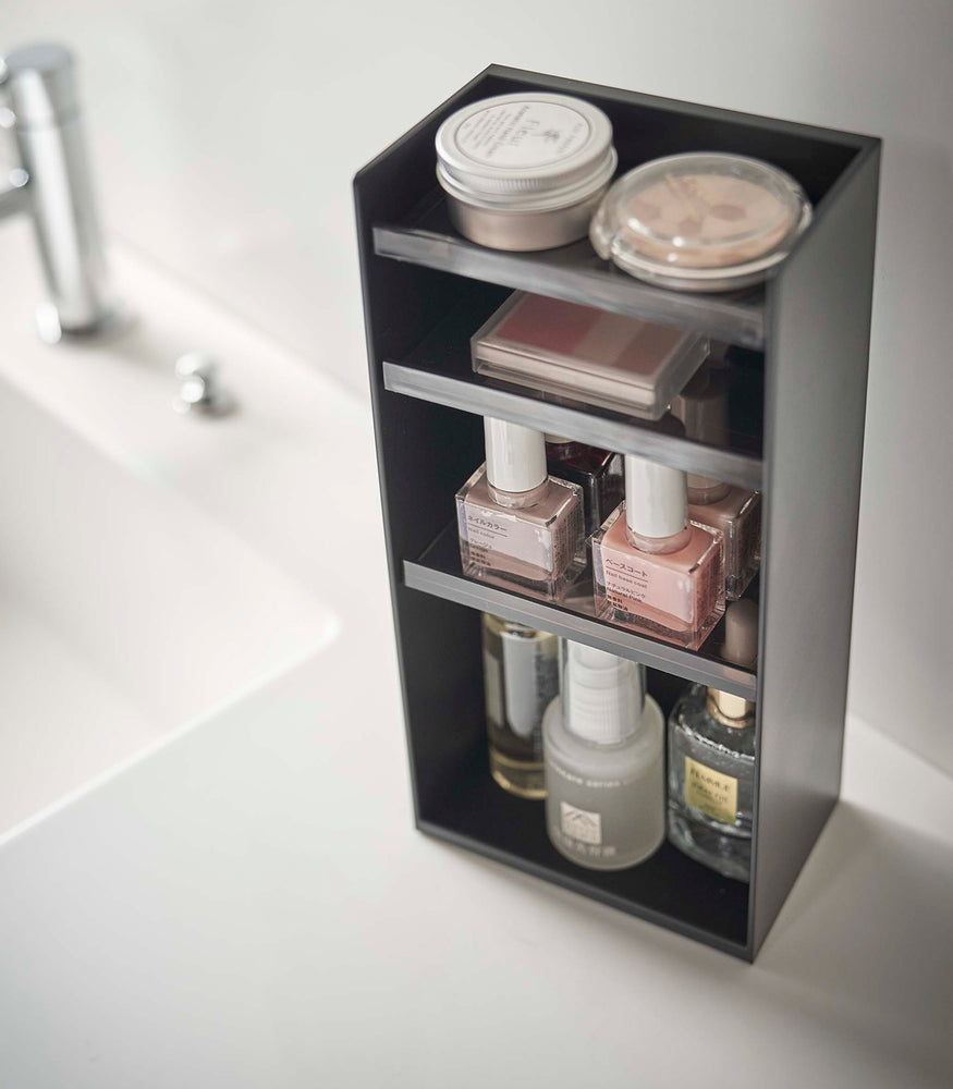 View 33 - Angled view of a black rectangular cosmetics organizer sitting on a white bathroom counter. The organizer has three black transparent shelves with upward facing lips to prevent the products from falling-out.
