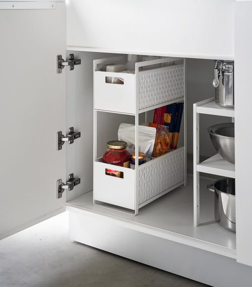 A white two-tier storage bin is shown in a lower cabinet. The upper level holds cleaning supplies, the bottom level holds pantry items.