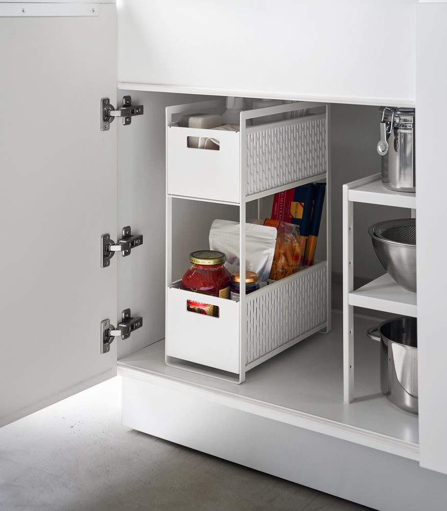 View 2 - A white two-tier storage bin is shown in a lower cabinet. The upper level holds cleaning supplies, the bottom level holds pantry items.