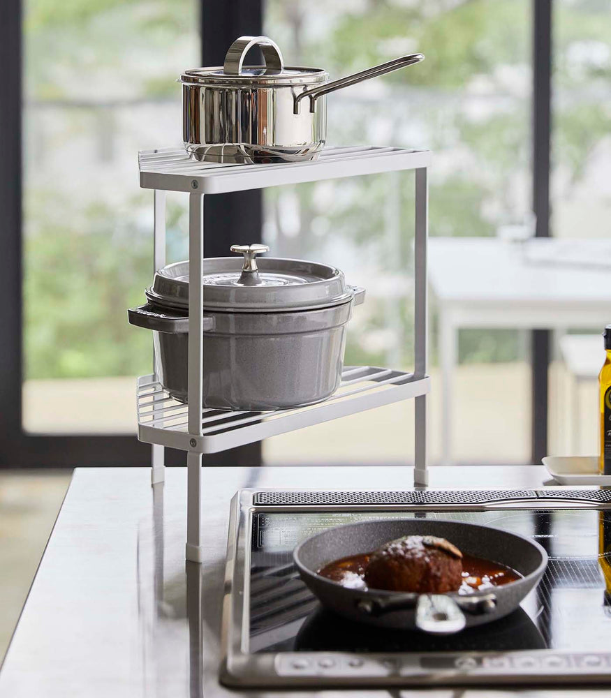 View 2 - White Two-Tier Corner Riser holding pot and saucepan next to stovetop by Yamazaki Home.