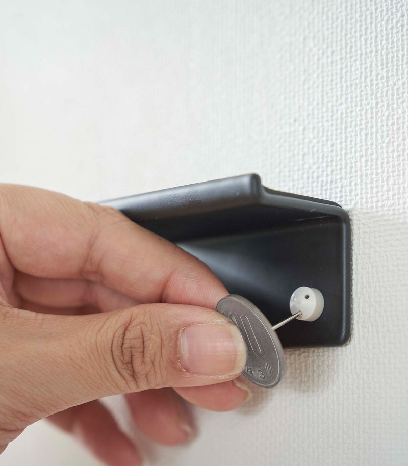 A hand is shown using a coin to secure nails into a wall, installing a black wall-mounted phone/tablet holder.