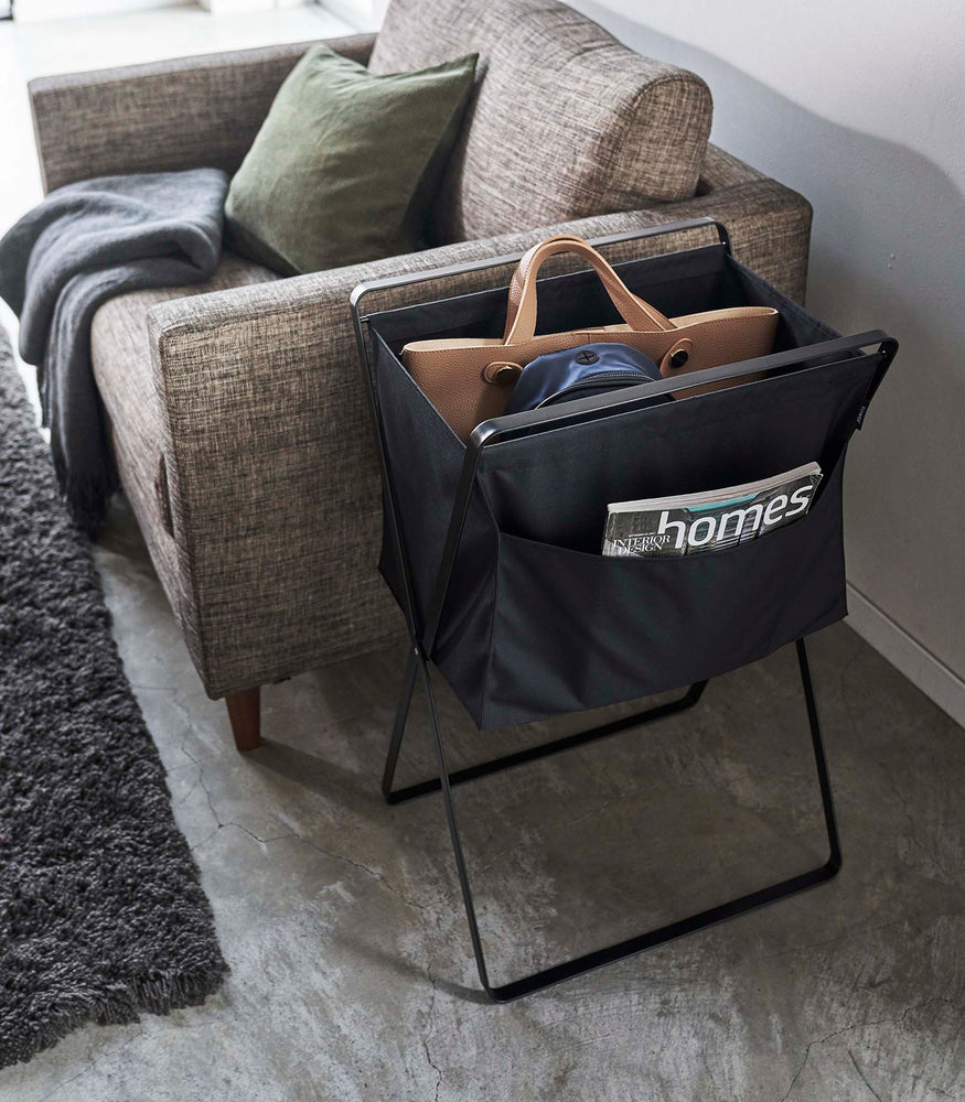 View 28 - A black canvas hamper holding a purse and backpack. Magazines are seen peeking out of a side-pocket. A brown couch with a green throw pillow and gray rug can be seen in the background.