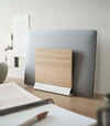 A small light-colored wood laptop stand with a white metal base stores a closed laptop on a wooden desk.
