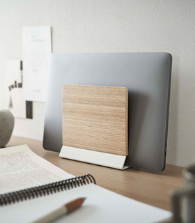 A small light-colored wood laptop stand with a white metal base stores a closed laptop on a wooden desk. view 2