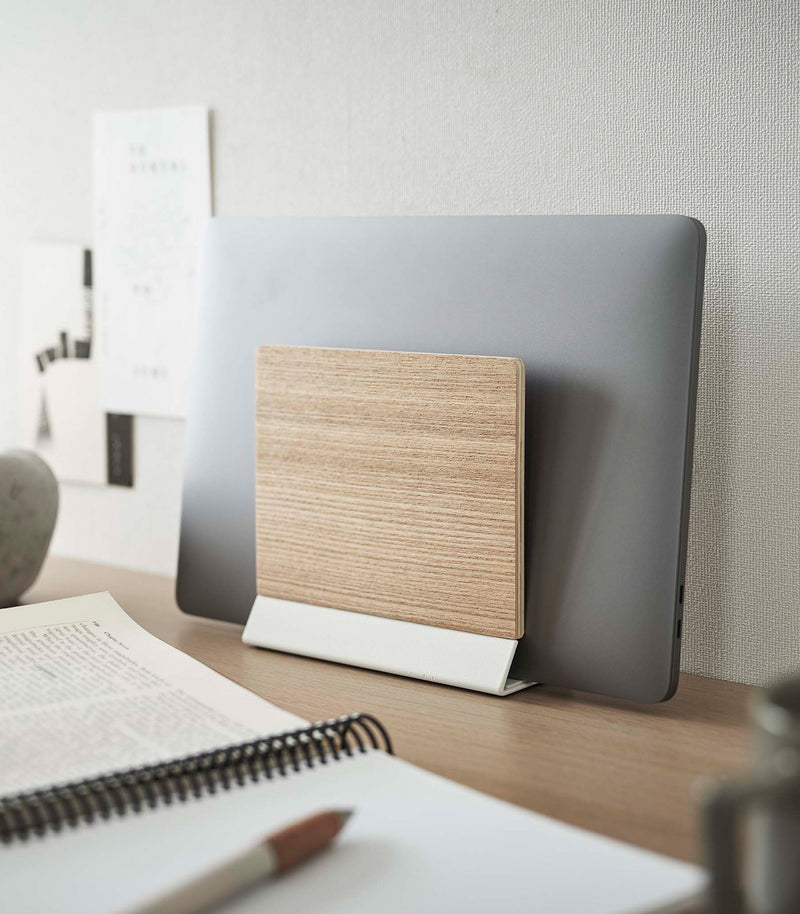 A small light-colored wood laptop stand with a white metal base stores a closed laptop on a wooden desk.