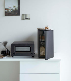 A vertical black metal breadbox is seen on a white kitchen counter next to a black microwave oven and a glass drip coffee pot with a dark liquid inside. view 24