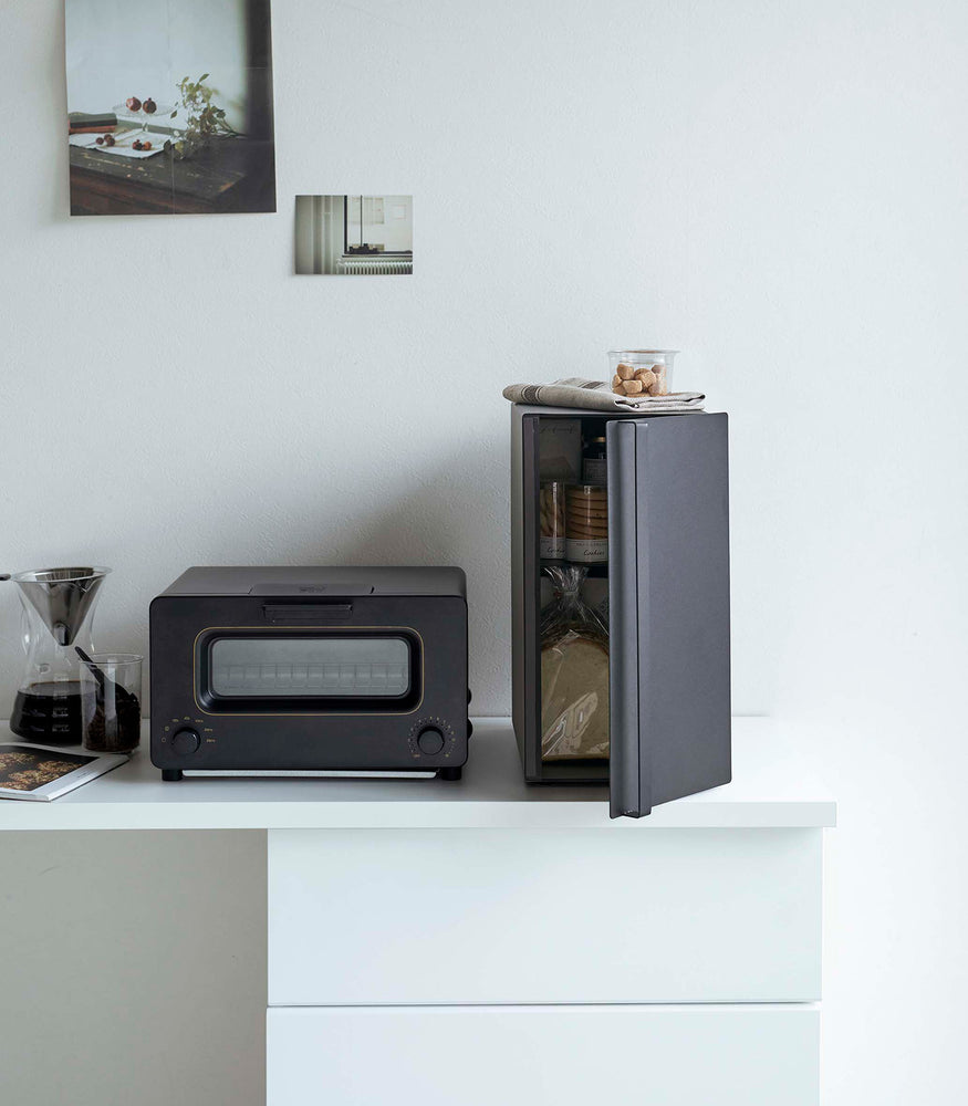 View 24 - A vertical black metal breadbox is seen on a white kitchen counter next to a black microwave oven and a glass drip coffee pot with a dark liquid inside.