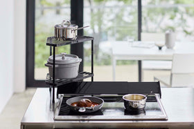 Front view of Black Two-Tier Corner Riser holding pot and saucepan next to stovetop by Yamazaki Home. view 9