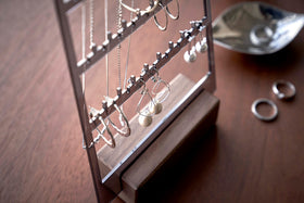A detailed view of an acrylic translucent mauve earring holder with a rectangular wooden base on a dark wood dresser. The acrylic holder has upward pointed hooks and slots placed in an interchangeable pattern. Hanging from the hooks are chained necklaces, and in the slots are various earrings. view 11
