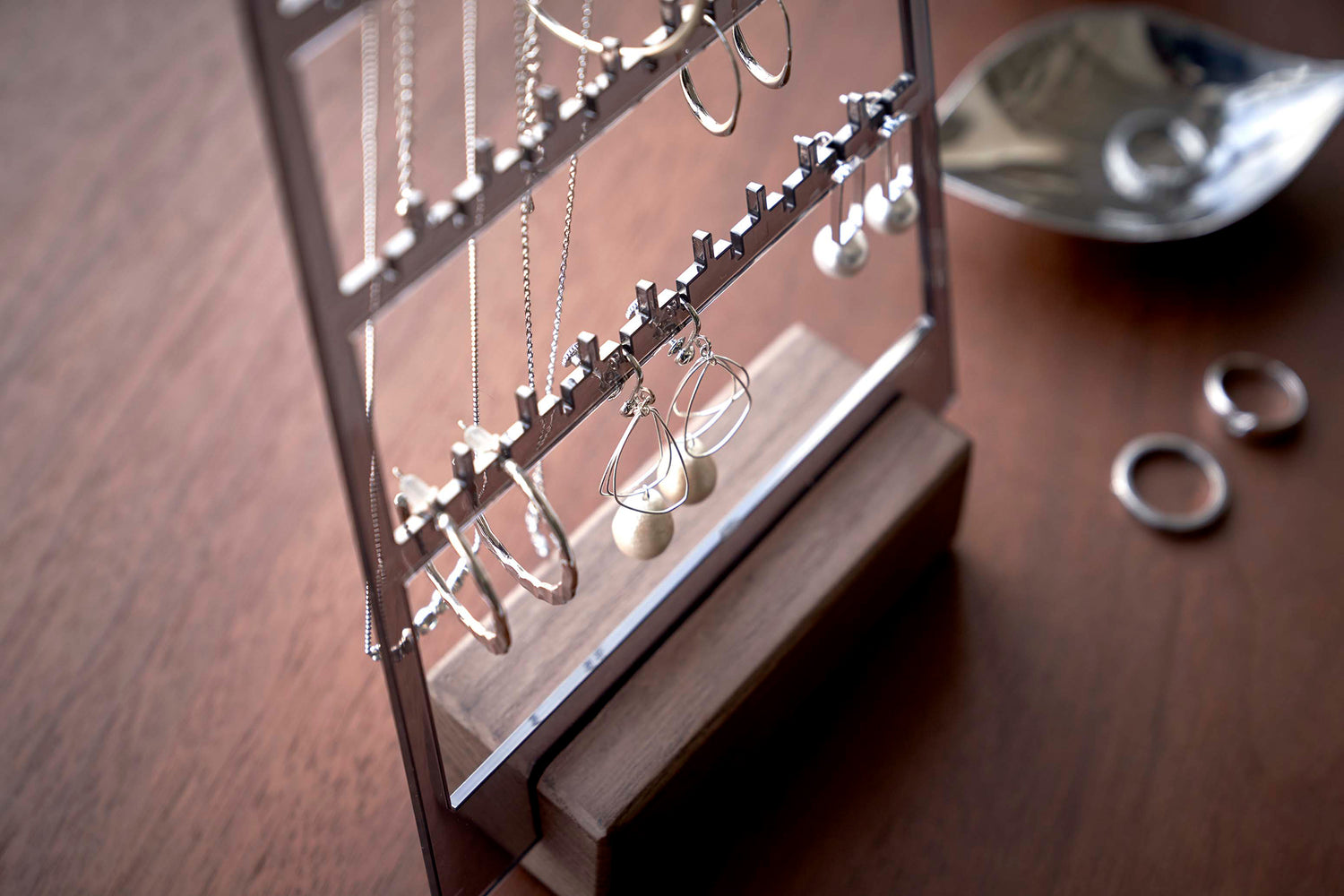View 11 - A detailed view of an acrylic translucent mauve earring holder with a rectangular wooden base on a dark wood dresser. The acrylic holder has upward pointed hooks and slots placed in an interchangeable pattern. Hanging from the hooks are chained necklaces, and in the slots are various earrings.