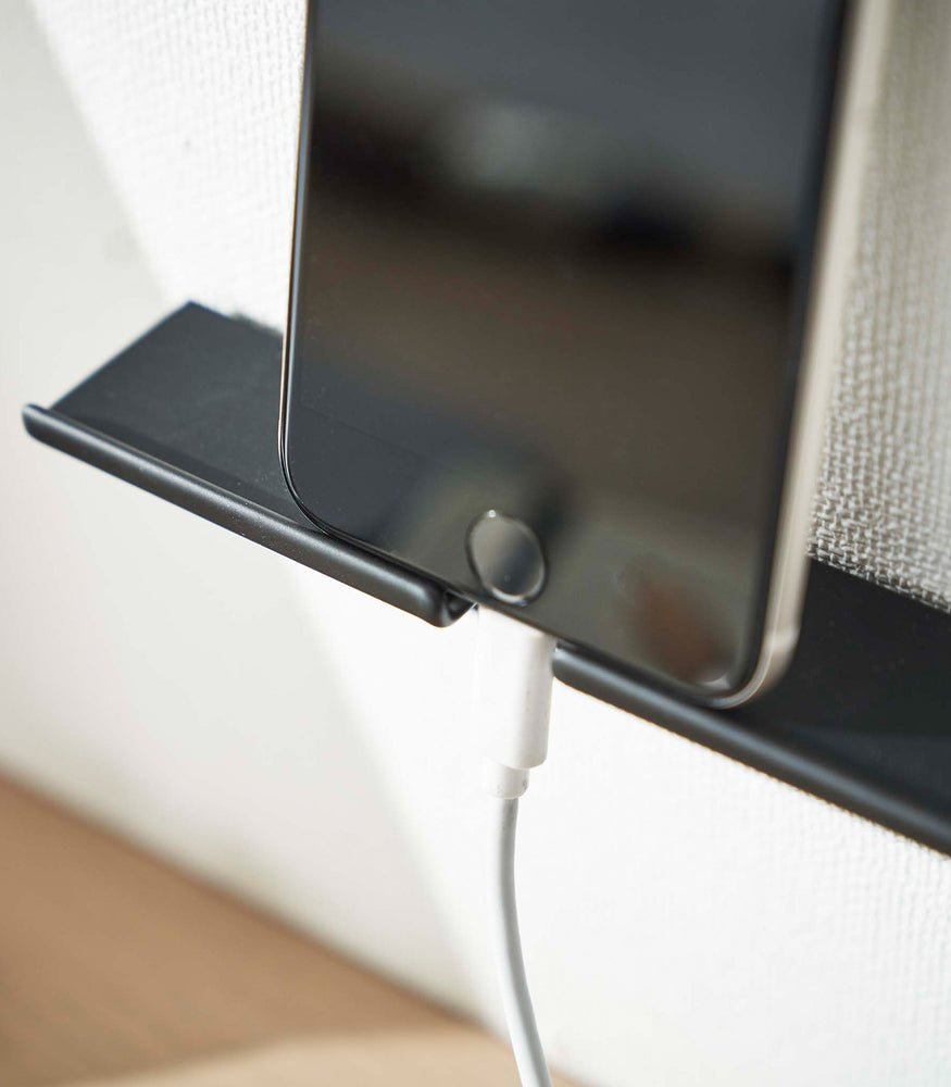 View 13 - An iPhone securely mounted on a wall, showcasing its screen while positioned vertically. The charger cord is neatly threaded through a designated opening in the mount, ensuring a streamlined appearance