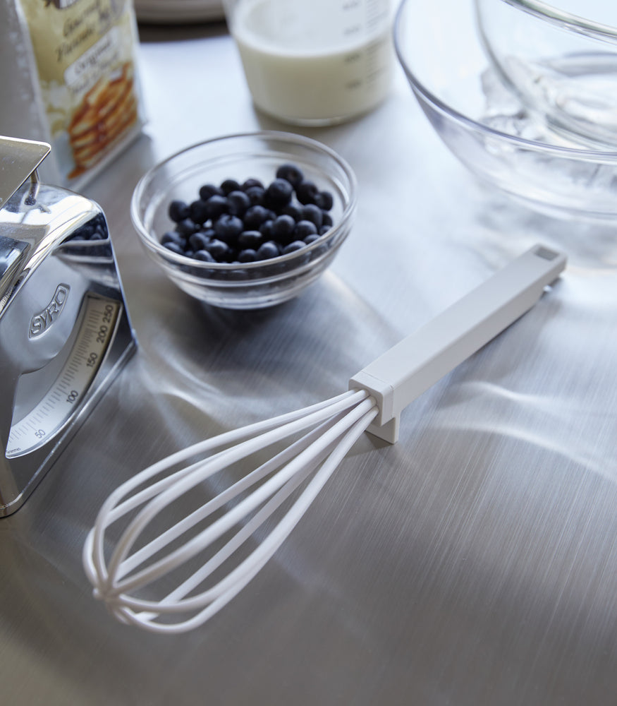 View 2 - A white Yamazaki Floating whisk resting on a table next to blueberries.
