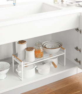 Front right diagonal view of white wired organizer rack holding pots, jars, and bowls under the sink. view 6
