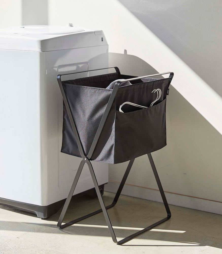View 23 - A black laundry hamper with black metal legs is angled in front of a washing machine. Wired hangers are poking out of a pocket in the front of the hamper and a towel is seen inside. A washing machine and bathtub are visible in the background.