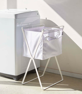 A white canvas hamper with white metal legs is positioned next to a washing machine. Wired hangers are seen poking out of a side pocket on the hamper. view 3