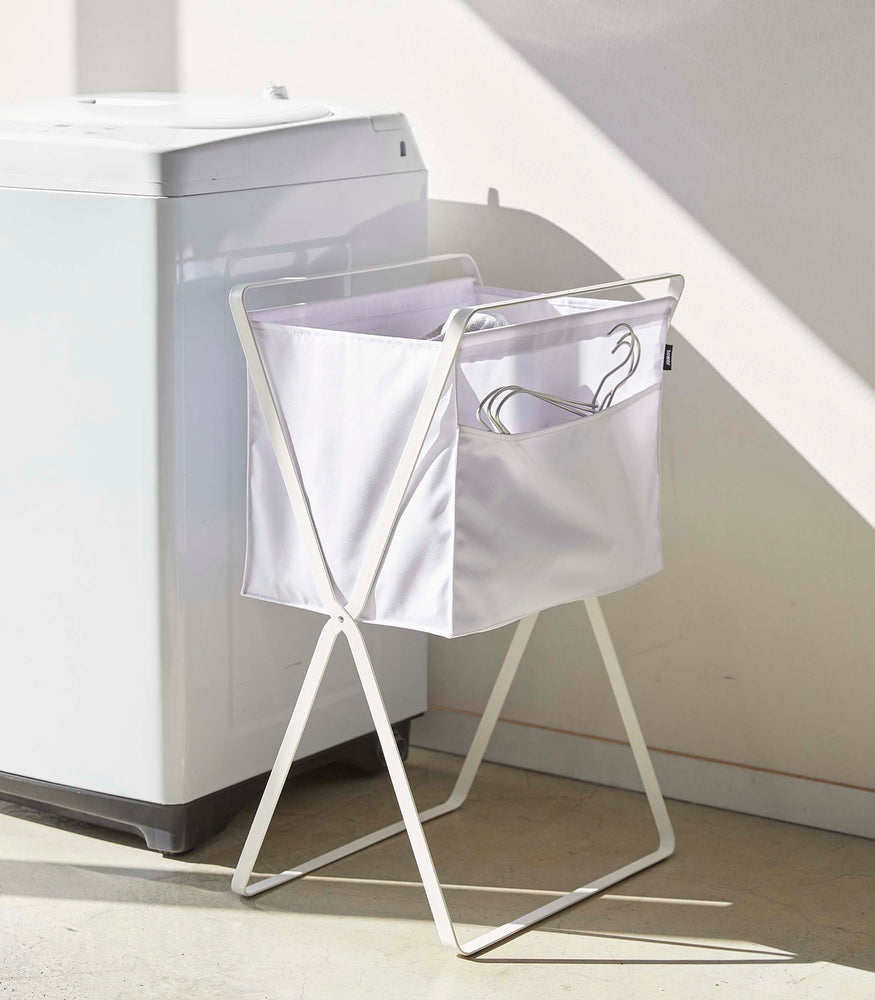 View 15 - A white canvas hamper with white metal legs is positioned next to a washing machine. Wired hangers are seen poking out of a side pocket on the hamper.