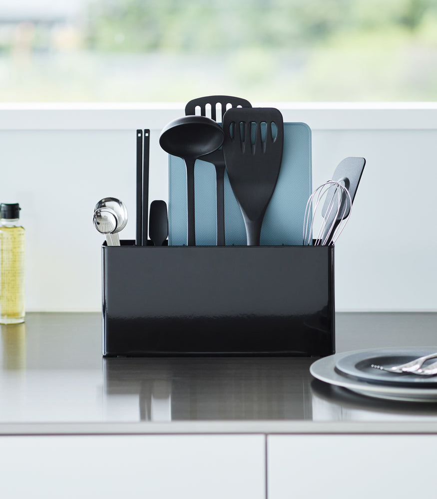 View 12 - Black Utensil & Thin Cutting Board Holder by Yamazaki Home on a kitchen counter, holding various utensils and a cutting board.