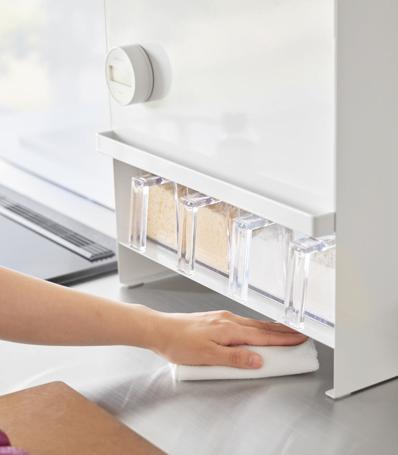 Image of a hand holding a duster and cleaning the countertop area under the Concealable Spice Rack by Yamazaki Home in white.