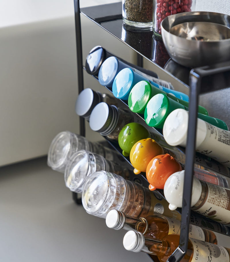 Close-up of a Black Four-Tier Slim Spice Rack by Yamazaki Home with spices, oils, and cooking utensils on a kitchen counter.