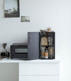 A vertical black metal breadbox is seen on a white kitchen counter next to a black microwave oven and a glass drip coffee pot with a dark liquid inside. view 26