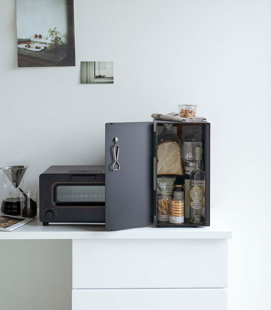 View 26 - A vertical black metal breadbox is seen on a white kitchen counter next to a black microwave oven and a glass drip coffee pot with a dark liquid inside.
