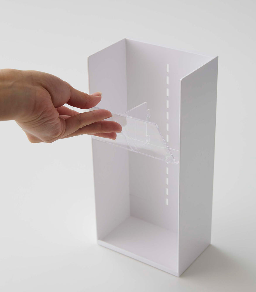 View 18 - A male hand pulls a deep transparent tray to adjust the location in a cosmetics organizer. It is a white resin rectangular cosmetics holder with an open face and top. The removable tray acts as a display and sits diagonally for easy visibility of the products held. Small evenly distributed rectangular cut-outs go down the middle of the organizer, allowing the trays locations to be adjusted. The bottom of the organizer has a slight upward pointing lip.
