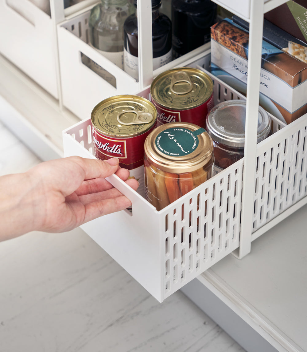 Three two-tier white storage bins are displayed in a lower cabinet. Both levels of each hold various food products. A model is sliding the lower level of one out to show its accessibility.