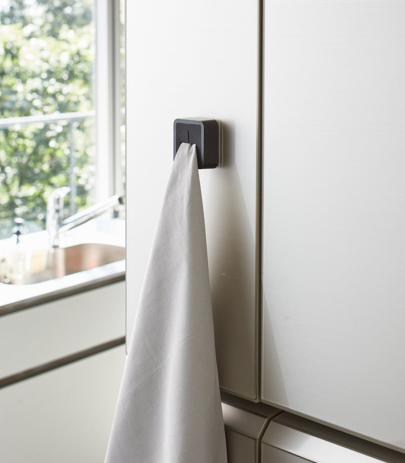 Black Yamazaki Home Traceless Adhesive Towel Holder attached to a cabinet with a towel inserted